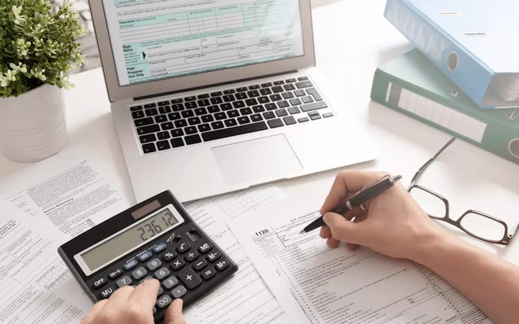 As mãos de uma pessoa sobre uma mesa de trabalho fazendo contas com uma calculadora e um notebook